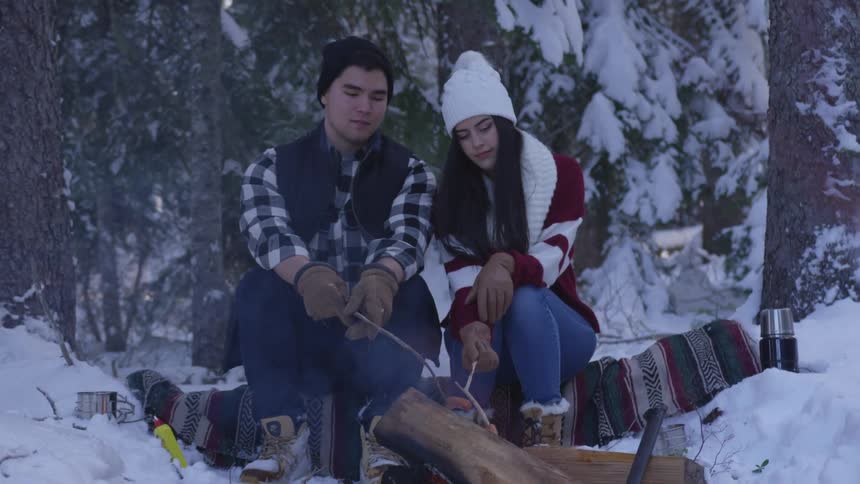 A man and woman sit outside in the snow and roast hot dogs over a fire