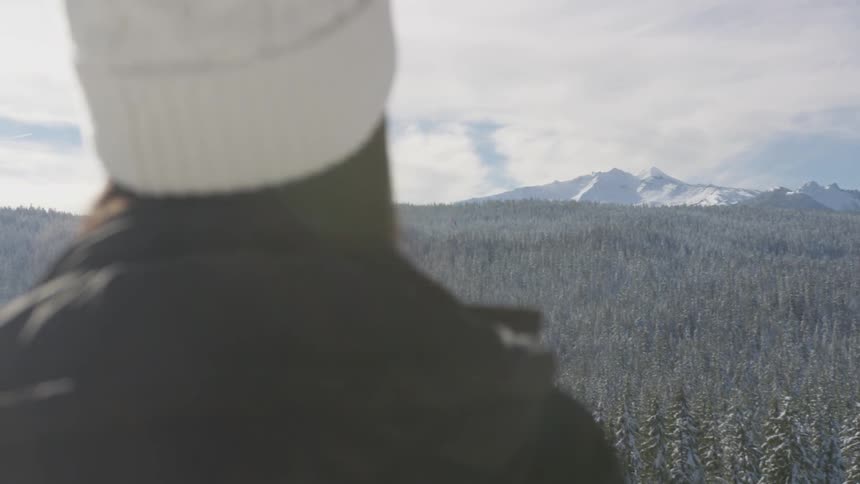 A woman stands on a cliff looking at the snow covered forest and mountain