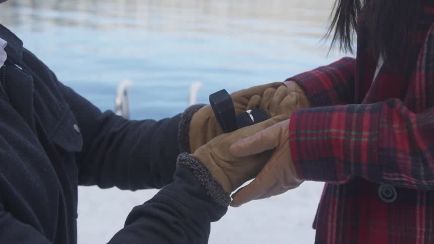A man on his knee is holding a black box with a diamond engagement ring and his fiance is holding his hands outside in the snow