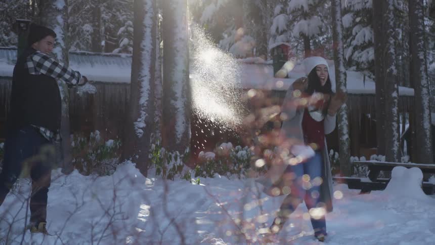A young man throws a snowball at a young woman in front of a cabin in a forest
