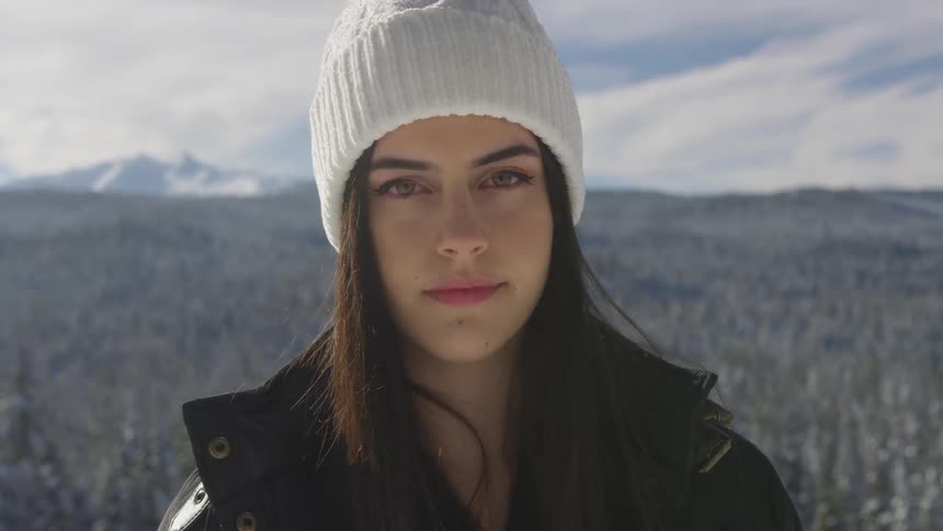 A dark haired female in a winter hat stands in front of a field with mountains in the background