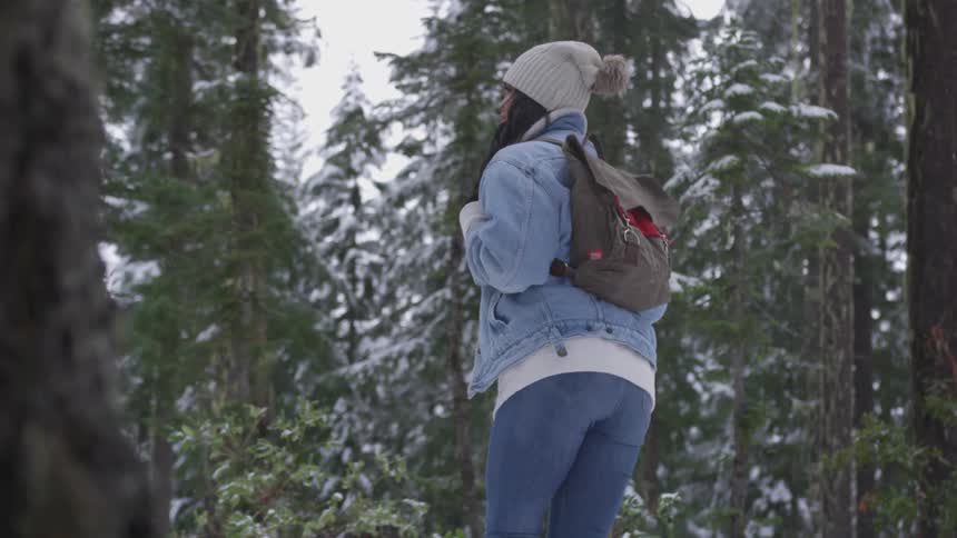 A black woman with a backpack hat and jean jacket hikes through the snowy woods