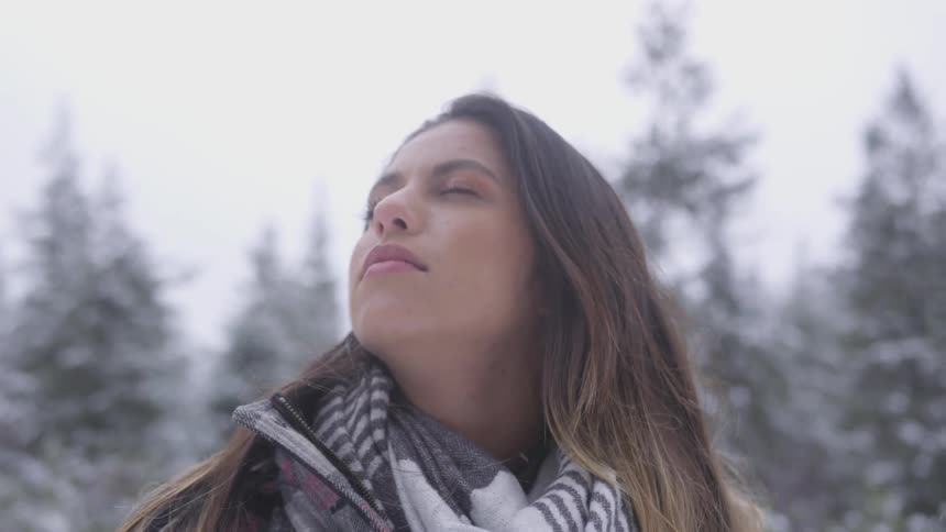 Brunette woman closes eyes and keeps her head up while the snow falls slowly in the forest in the daytime