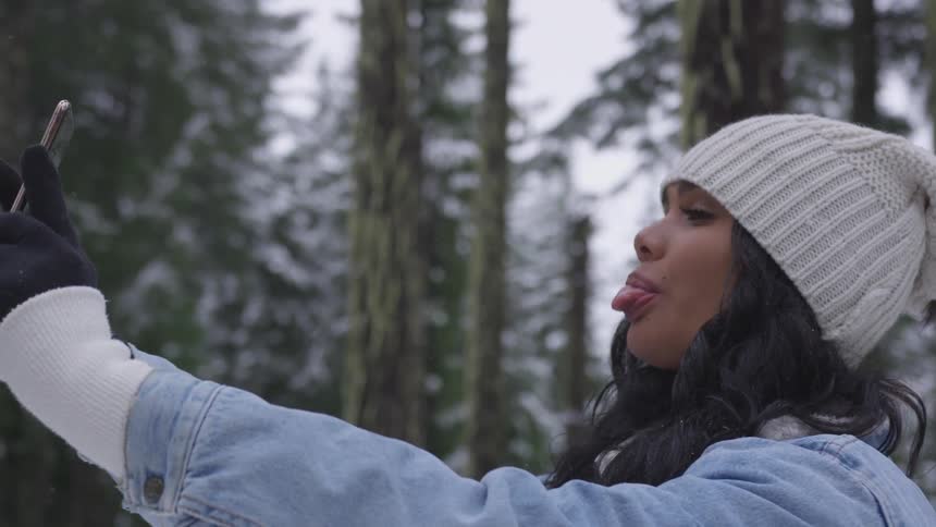 Brunette woman wearing wooly hat puts out her tongue and takes a selfie in the forest during daylight