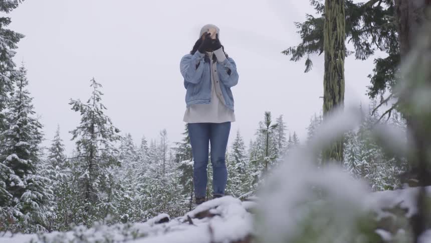 Woman in the winter clothes stands in the forest and makes photos with her phone