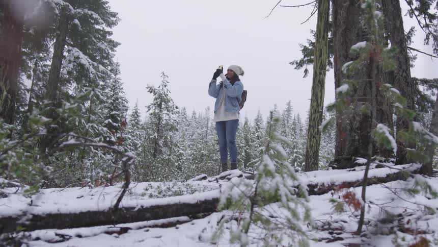 Woman stands in snow covered forest taking pictures with her mobile phone
