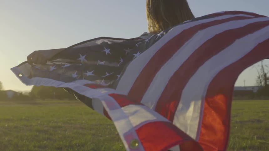 A female teenager stands in an open field holding an american flag behind her which blows in the wind