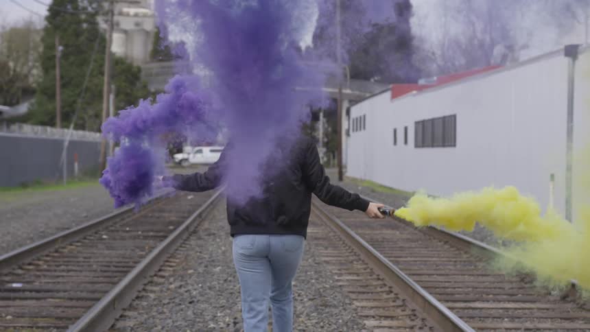 A woman is holding colored smoke signals while walking down a set of train tracks