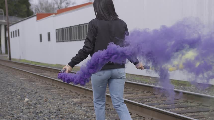 A hispanic woman walks near railroad tracks while holding two smoke grenades