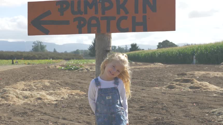 Little blonde girl stands in front of wooden sign with farmland background and smiles on a sunny day