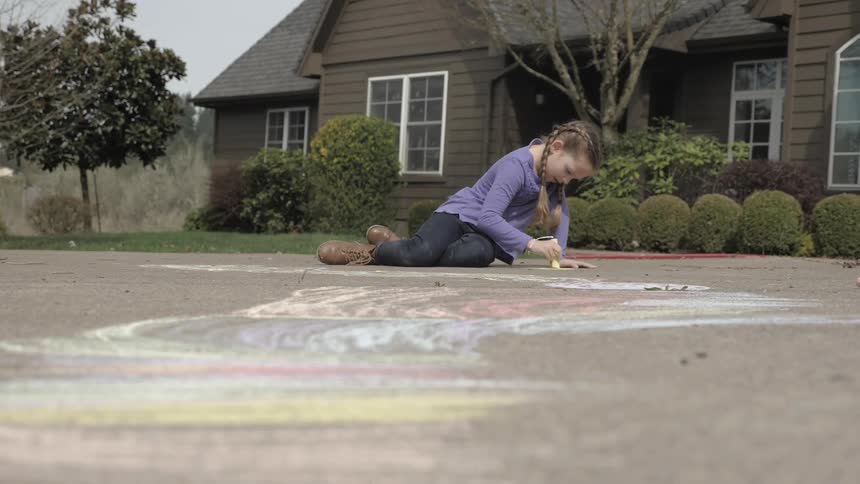 A young girl uses sidewalk chalk to draw on the street in front of a home with brown siding