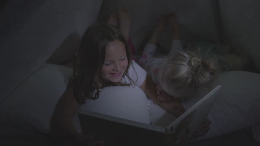 Two young white children lie in bed reading a book and giggling together