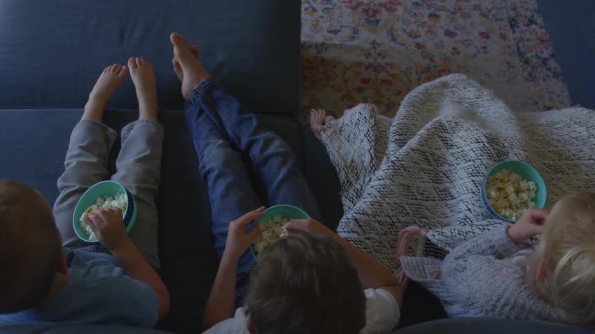 Three children sit on a couch eating popcorn