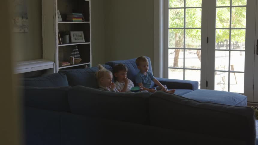 Children sitting on the blue sofa watch tv and eat nuts from a bowl in the daytime