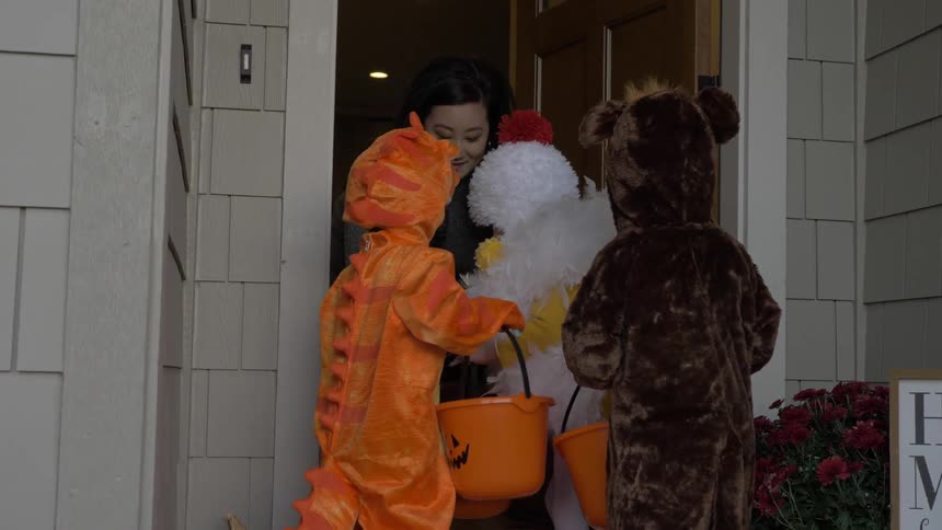 Three youngsters dressed head to toe in colorful animal and dinosaur costumes stand before a door answered by a smiling woman who begins to provide them with candy in the orange plastic trick or treating buckets that they are each holding