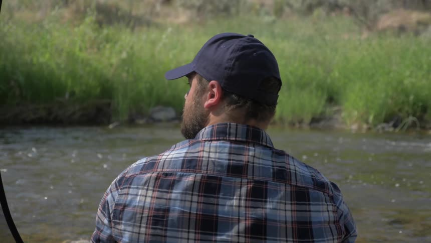 A white man wearing a ball cap sits on the side of a river and watches the water go by