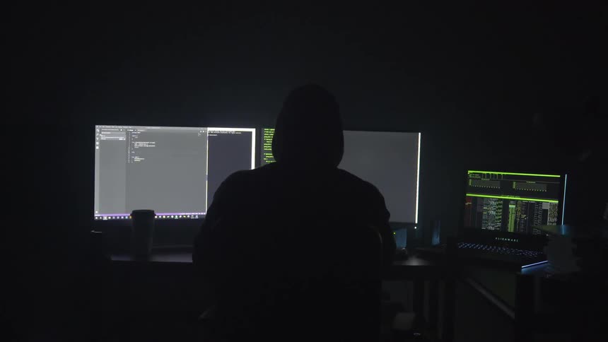 A person working from three monitors on a desk in a darkened room