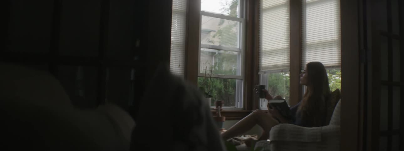 A girl sits down in a chair besides windows with a book and coffee
