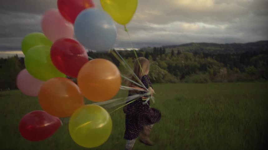 A little girl runs through a field while holding many balloons