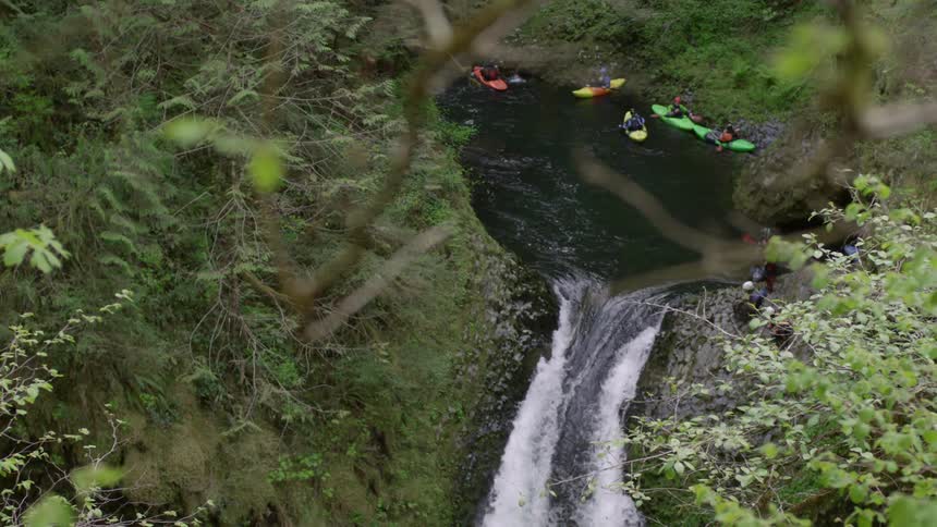 Five kayakers rest by the river bank opposite a waterfall contemplating running the rapid below