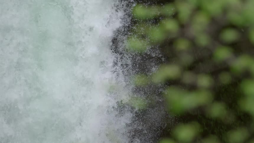 Tight slow motion shot of a huge waterfall cascading to the valley floor below with green ferns alongside the edge of the frame