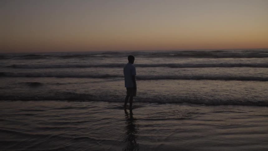 A boy stands on the sand while ocean waves crash against his feet