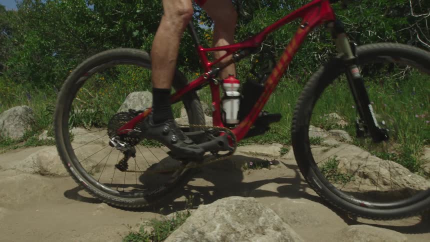 A person rides their bicycle over rocks and sand near green grass and bushes