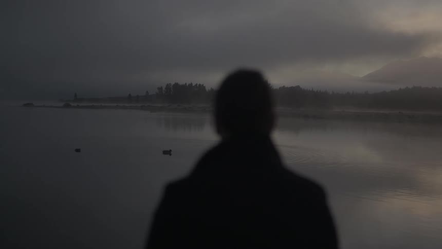 Man stands alone by a foggy lake at dusk
