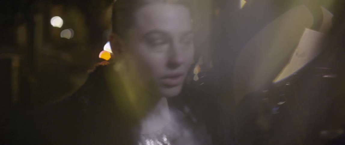 A woman wearing a shiny gold dress and fur coat walking and talking then stopping to put her hand to her forehead