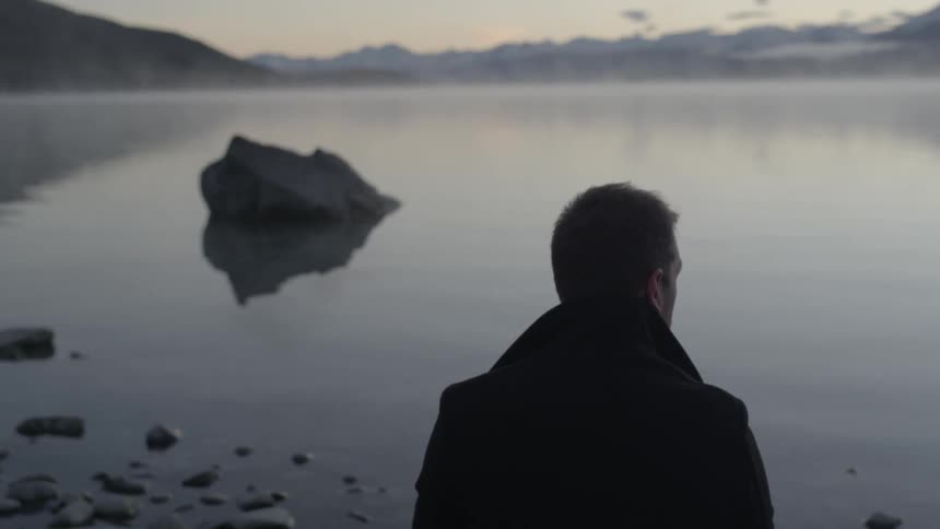 A man gazing over a still lake with mountains in the background