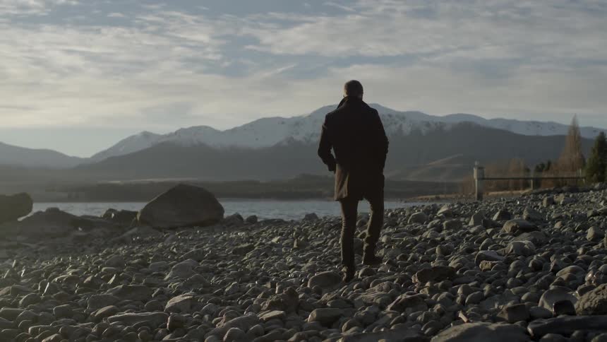 A man walks on a rocky beach towards a large body of water with mountains in the distance