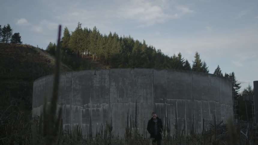 A man walking away from a round cement structure next to a tree-lined hill