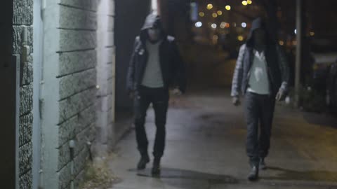 A man walks down a street at night while being followed by two men in masks