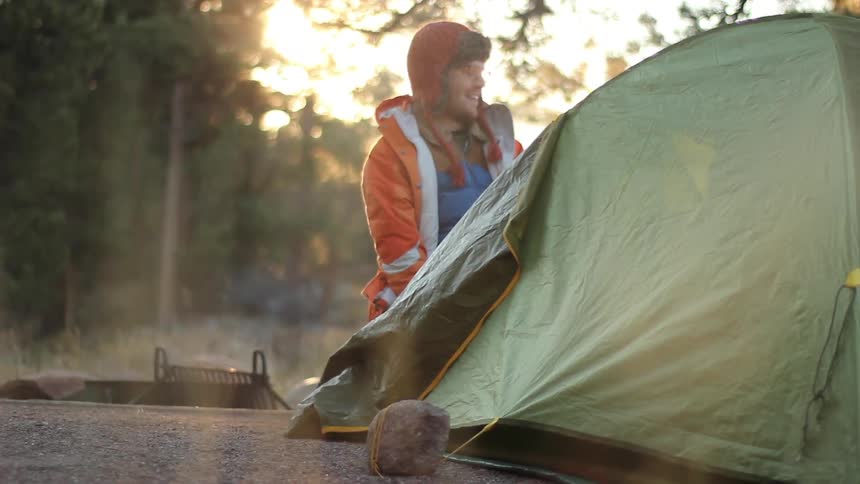 Man squats near tent while a campfire smokes in the background