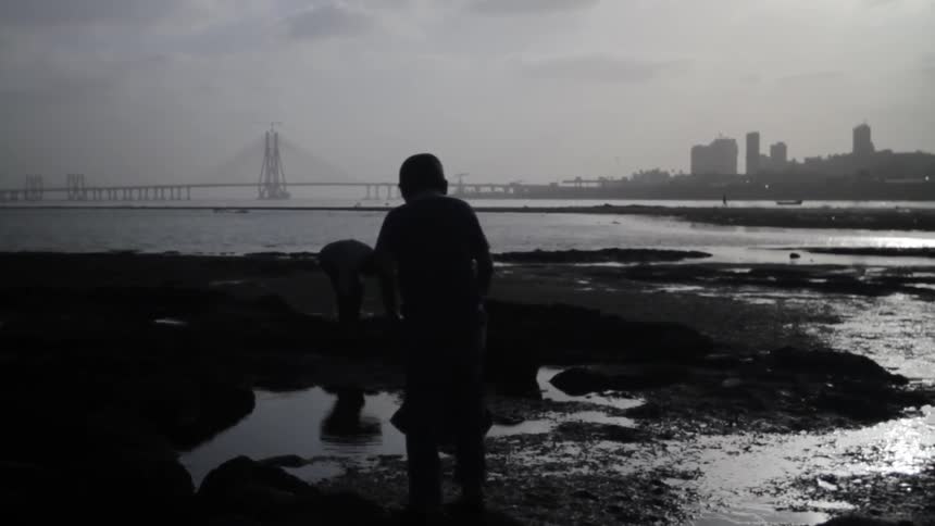 Two young boys playing on sand near puddles next to the shoreline with a view of a bridge and city skyline in the background