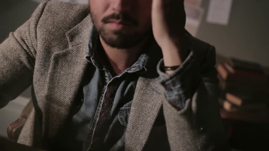 A man in a sports coat and glasses sitting at a desk shows frustration at paperwork