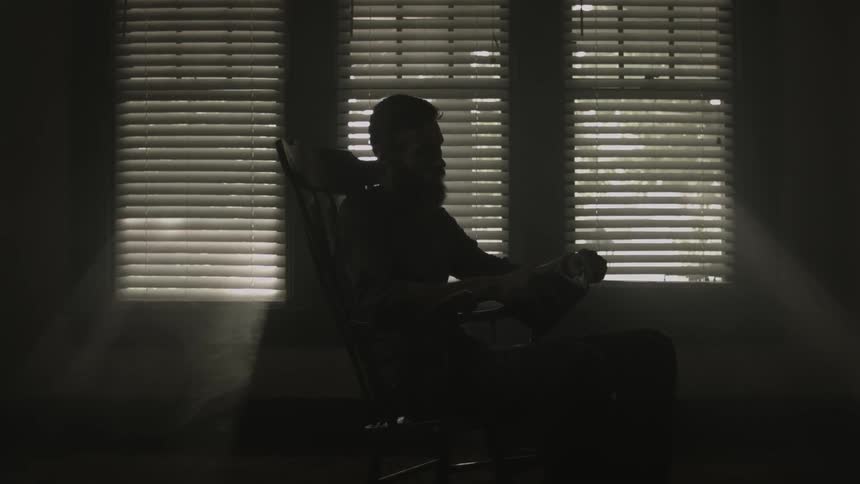 A bearded man sitting in the shadows on a rocking chair in front of windows