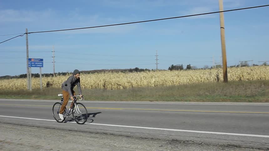 A man bicycles down a road passing a corn field