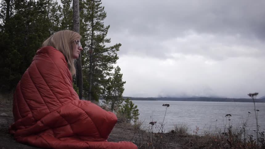 A woman wrapped in a sleeping bag looks out over a lake