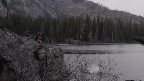 People sitting on a cliff as a man jumps off the cliff and into a body of water that is surrounded by trees and has mountains in the background