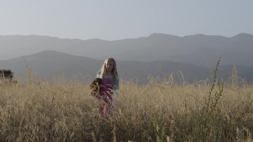 A young blonde girl carrying a doll and walking through tall grass with mountains in the background