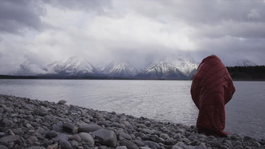 Blonde woman wrapped in the red sleeping bag stands by the lakeside with the snow capped mountain ranges