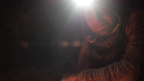 A man wearing a miner's light on his head adjusting something on the ground and then looking towards the camera while smiling and speaking