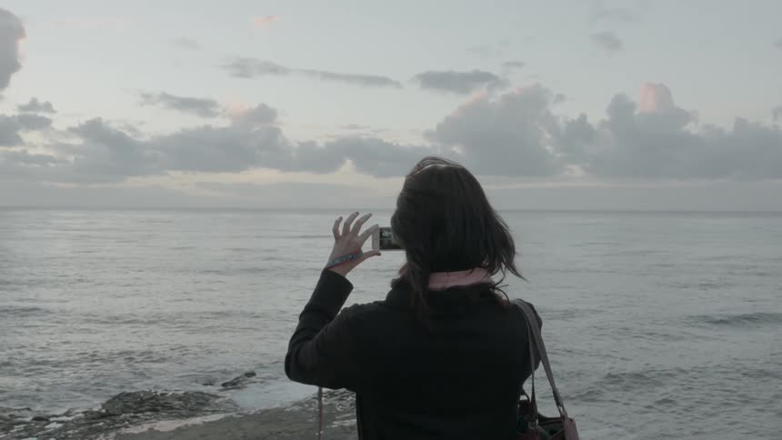 A woman uses an iphone to take a picture near the beach