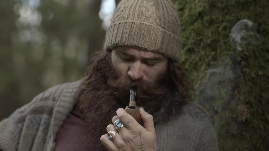 Bearded man smokes his pipe against a tree