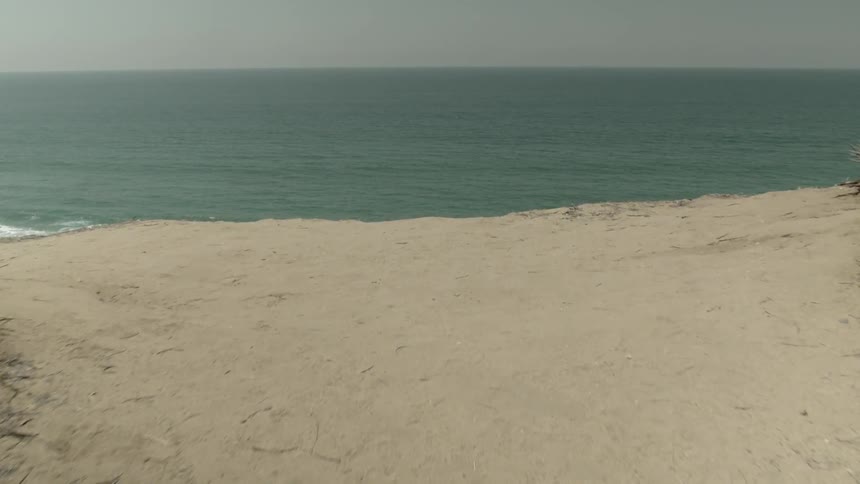 First person view of walking to the edge of a sandy cliff and looking down at the rocky shoreline as waves crash on shore