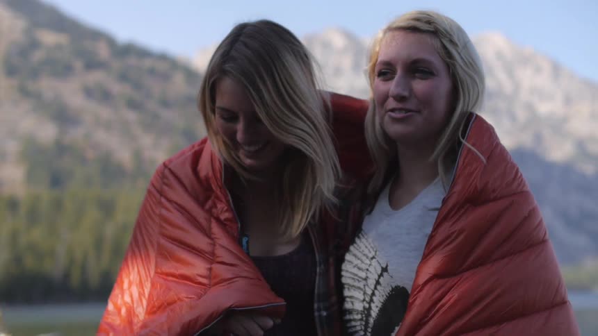 Two blonde women covering with a sleeping bag poses for the camera with mountains in the background