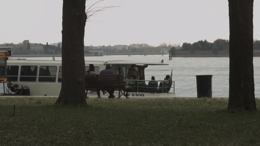 People sitting on a bench as a tourist boat pulls away