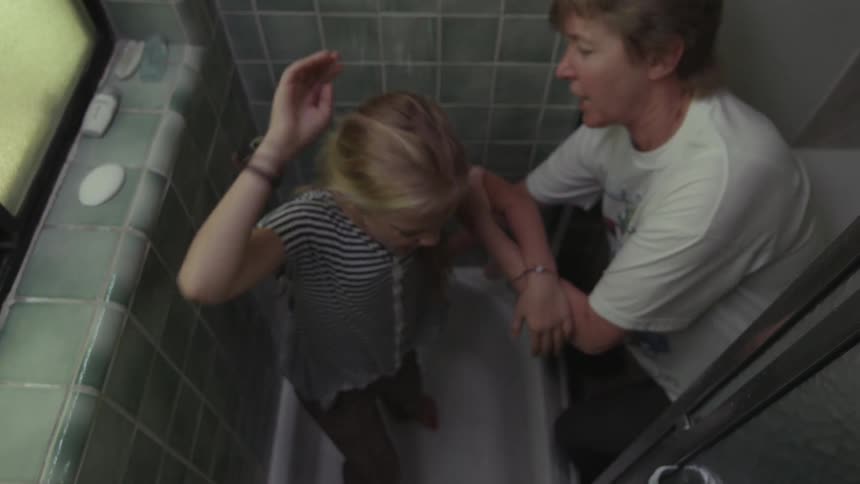 An older woman forcefully puts a little girl in a shower fully clothed