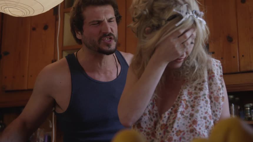A man holding a beer bottle in one hand while he yells at a blonde woman who turns away and cowers to the floor as his verbal abuse escalates